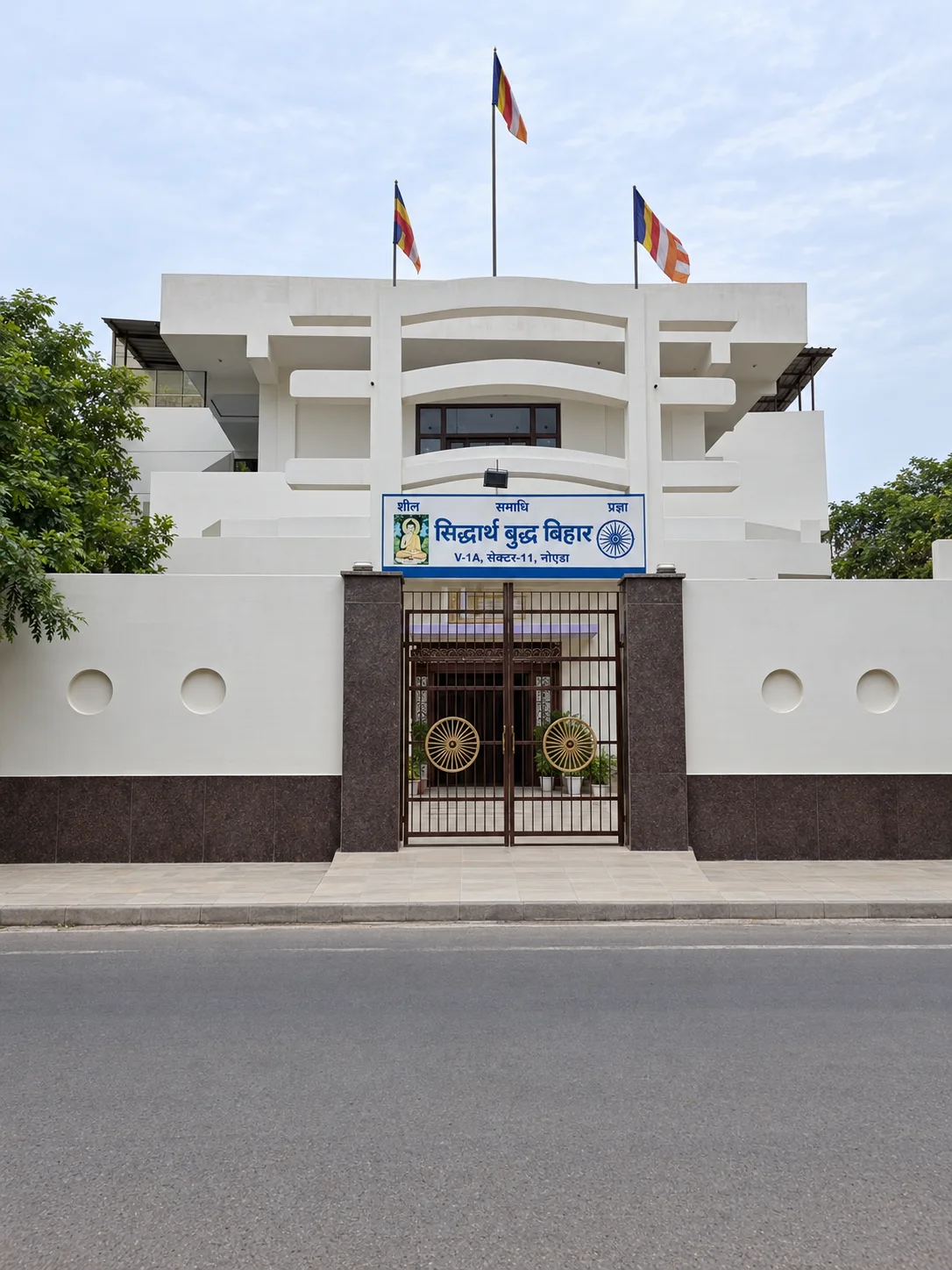 Front entrance and white façade of Siddharth Buddh Vihar in Noida, with Buddhist flags above the building.